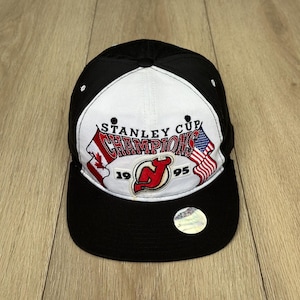 Könnte beinhalten: Schwarz-weiße Baseballkappe mit dem Text "Stanley Cup Champions" und den Flaggen von Kanada und den Vereinigten Staaten. Die Kappe zeigt das Logo der New Jersey Devils und das Jahr 1995.