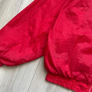 May include: A close-up of a bright red nylon jacket. The jacket has a zipper closure and elastic cuffs and waistband. The fabric has a slight sheen, and the jacket appears to be lightweight.