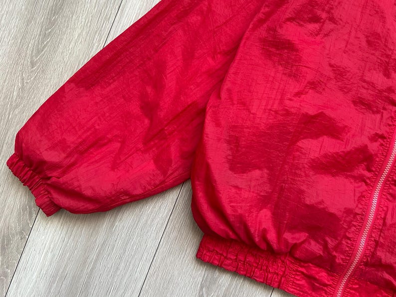 May include: A close-up of a bright red nylon jacket. The jacket has a zipper closure and elastic cuffs and waistband. The fabric has a slight sheen, and the jacket appears to be lightweight.