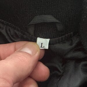 May include: Close-up of a black garment with a white label indicating size "L". The label is held by a hand, showing the garment's interior lining. The fabric appears to be a dark, possibly quilted material.