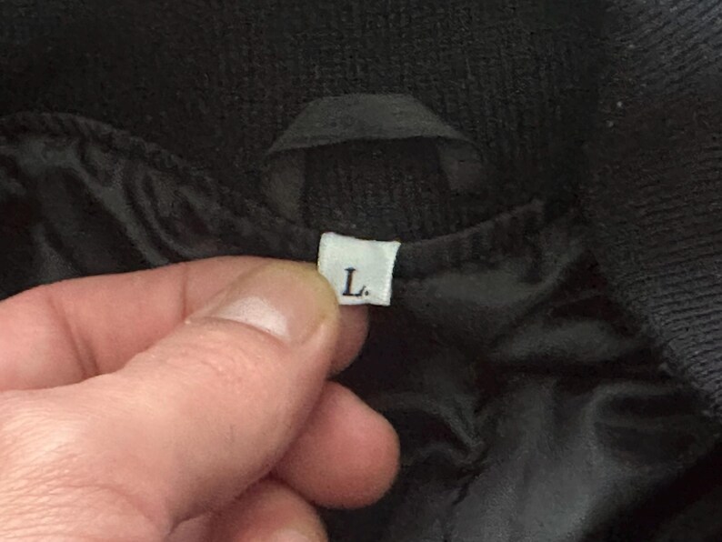 May include: Close-up of a black garment with a white label indicating size "L". The label is held by a hand, showing the garment's interior lining. The fabric appears to be a dark, possibly quilted material.