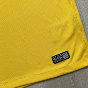 Pu&ograve; includere: Primo piano di un tessuto giallo brillante con una superficie strutturata. Un'etichetta nera con la parola "AUTHENTIC" e altro testo &egrave; cucita sul tessuto. L'etichetta ha anche un piccolo logo Nike.