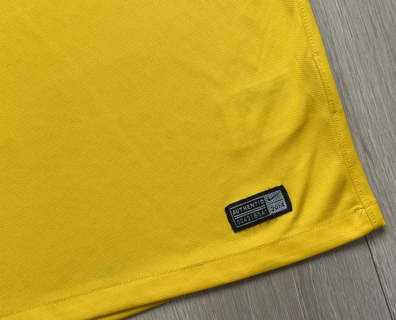 Pu&ograve; includere: Primo piano di un tessuto giallo brillante con una superficie strutturata. Un'etichetta nera con la parola "AUTHENTIC" e altro testo &egrave; cucita sul tessuto. L'etichetta ha anche un piccolo logo Nike.