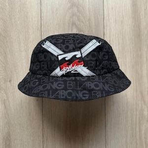 May include: Black bucket hat with the word "Billabong" repeated in a pattern. The hat features a logo with a red and black flame design, crossed white lines, and the brand name in white script.
