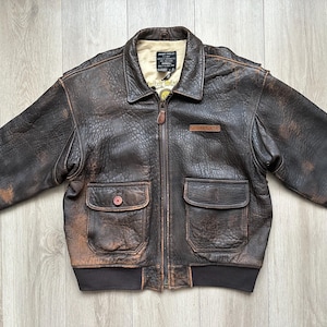 May include: A dark brown distressed leather bomber jacket featuring a pointed collar, a front zip fastening, and two large flap pockets. The interior lining is cream-coloured, and a black label with white text is visible at the neckline.