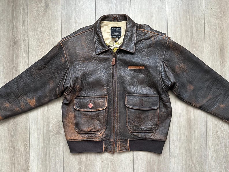 May include: A dark brown distressed leather bomber jacket featuring a pointed collar, a front zip fastening, and two large flap pockets. The interior lining is cream-coloured, and a black label with white text is visible at the neckline.
