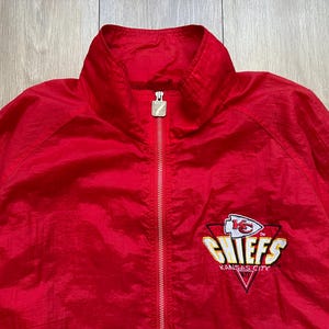 May include: A red zip-up jacket featuring the Kansas City Chiefs logo on the chest. The logo includes a white arrowhead, the word "CHIEFS" in gold, and the words "KANSAS CITY" in white. The jacket has a stand-up collar.