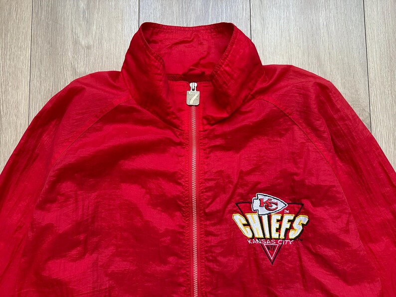 May include: A red zip-up jacket featuring the Kansas City Chiefs logo on the chest. The logo includes a white arrowhead, the word "CHIEFS" in gold, and the words "KANSAS CITY" in white. The jacket has a stand-up collar.