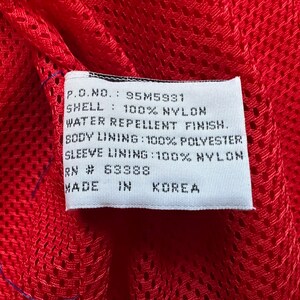 May include: Close-up of a white tag with black text against a red mesh fabric. The tag reads: P.O.NO.: 95M5931, SHELL: 100% NYLON, WATER REPELLENT FINISH, BODY LINING: 100% POLYESTER, SLEEVE LINING: 100% NYLON, RN # 63388, MADE IN KOREA.