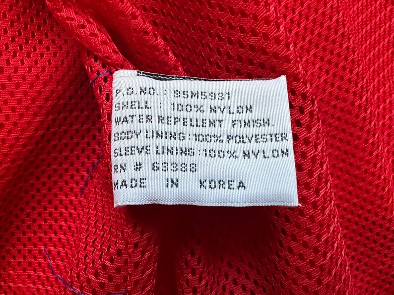 May include: Close-up of a white tag with black text against a red mesh fabric. The tag reads: P.O.NO.: 95M5931, SHELL: 100% NYLON, WATER REPELLENT FINISH, BODY LINING: 100% POLYESTER, SLEEVE LINING: 100% NYLON, RN # 63388, MADE IN KOREA.