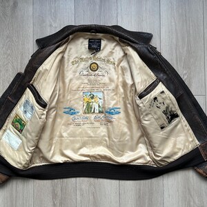 May include: A dark brown leather bomber jacket with a cream-coloured interior lining. The lining features a printed United States Navy Certificate of Service, a central illustration of sailors, and two additional rectangular graphic patches.