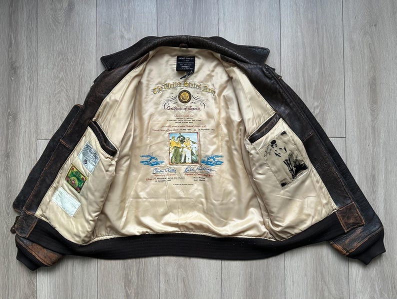 May include: A dark brown leather bomber jacket with a cream-coloured interior lining. The lining features a printed United States Navy Certificate of Service, a central illustration of sailors, and two additional rectangular graphic patches.