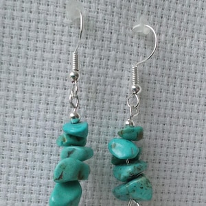 Peut inclure: Une paire de boucles d'oreilles en pierres turquoise. Chaque boucle d'oreille est composée d'un crochet et d'une chaîne argentés, avec une série de pierres de couleur turquoise, de forme irrégulière, enfilées ensemble. Les pierres ont une texture légèrement rugueuse.