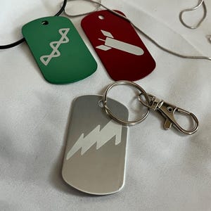 Puede incluir: Tres chapas de perro de metal con diferentes diseños. Una es verde con un diseño de escalera blanca, otra es roja con un diseño de cohete blanco y otra es plateada con un diseño de rayo blanco.