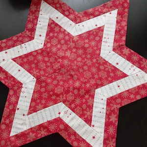 Könnte beinhalten: Ein roter und weißer sternförmiger Tischläufer mit einem Schneeflockenmuster und goldenen Akzenten. Der Läufer ist aus Stoff und hat eine gesteppte Textur.