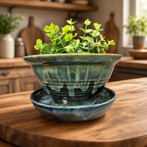 May include: A ceramic planter with a flared rim and a matching saucer, featuring a reactive glaze in shades of deep blue, teal, and green with a drip pattern. The planter has a visible drainage hole at the base and contains a green leafy plant.