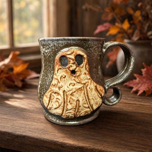 May include: A ceramic mug with a dark, mottled grey and brown glaze featuring a raised, cream-colored ghost figure on the front. The ghost has two dark, circular eyes and an open mouth, with intricate, swirling embossed patterns across its surface.