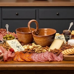 May include: A wooden charcuterie board with an assortment of meats, cheeses, nuts, olives, and crackers. Two brown ceramic bowls with handles are in the center. A jar of preserves and decorative serving spoons are also present.