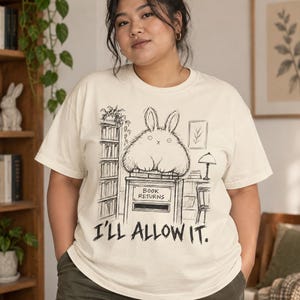 Peut inclure: Un t-shirt à manches courtes de couleur crème avec une illustration au trait noir représentant un lapin duveteux assis sur une boîte de retour de livres. Le motif inclut une bibliothèque, une lampe et le texte "BOOK RETURNS" et "I'LL ALLOW IT."