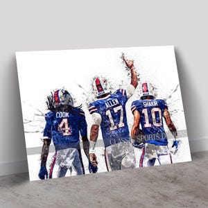 Peut inclure: Impression artistique de trois joueurs de football américain en uniformes bleus et blancs. Les joueurs sont vus de dos, avec leurs numéros et noms de maillot visibles. L'œuvre a un effet d'éclaboussure, avec le texte "CLASSIC SPORTS DESIGNS" en bas.