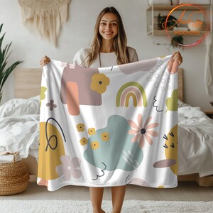 May include: A white fleece throw blanket with a colorful abstract pattern featuring rainbows, flowers, and geometric shapes. The pattern is in shades of pink, yellow, green, and blue.