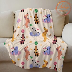 May include: A cream-colored fleece blanket with a repeating pattern of cartoon dachshunds in various colors, holding balls and hearts. The dachshunds are on a cream background with stars, hearts, and swirls.