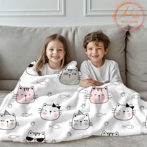 May include: Two children are sitting on a white couch with a white blanket with a black and pink cat pattern. The blanket has a repeating pattern of cats, fish, and paw prints. The children are smiling and looking at the camera.