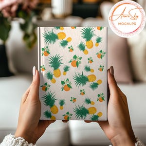 May include: A white notebook with a pineapple and orange pattern. The notebook is being held by a person's hands.