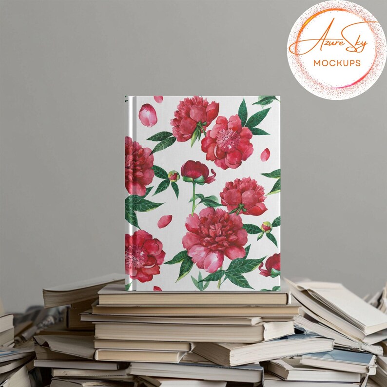 May include: A white notebook with a floral pattern of pink peonies and green leaves. The notebook is sitting on a stack of books.