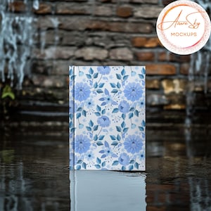 May include: A white notebook with a blue floral pattern on the cover. The notebook is sitting on a wet, gray surface.
