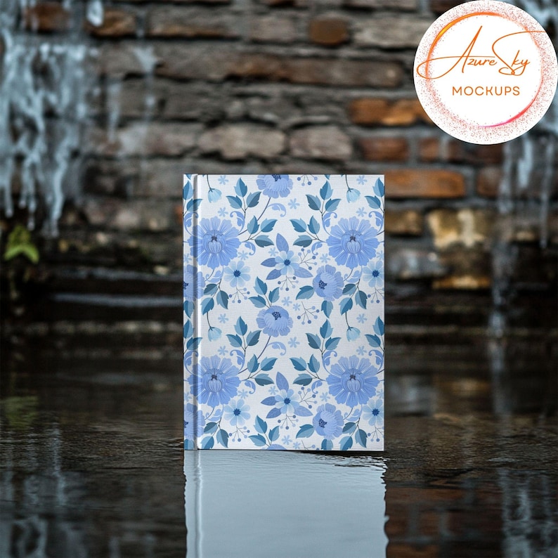 May include: A white notebook with a blue floral pattern on the cover. The notebook is sitting on a wet, gray surface.