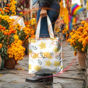May include: A white tote bag with a floral pattern of yellow flowers and green leaves. The bag has two white straps and is being held by a person wearing blue jeans.