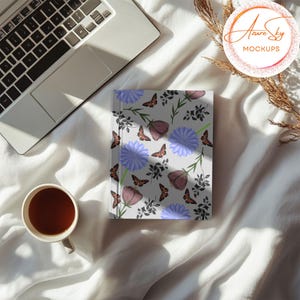 May include: A white notebook with a floral pattern featuring purple flowers, blue flowers, and brown butterflies. The notebook is on a white bedspread with a laptop and a cup of tea.