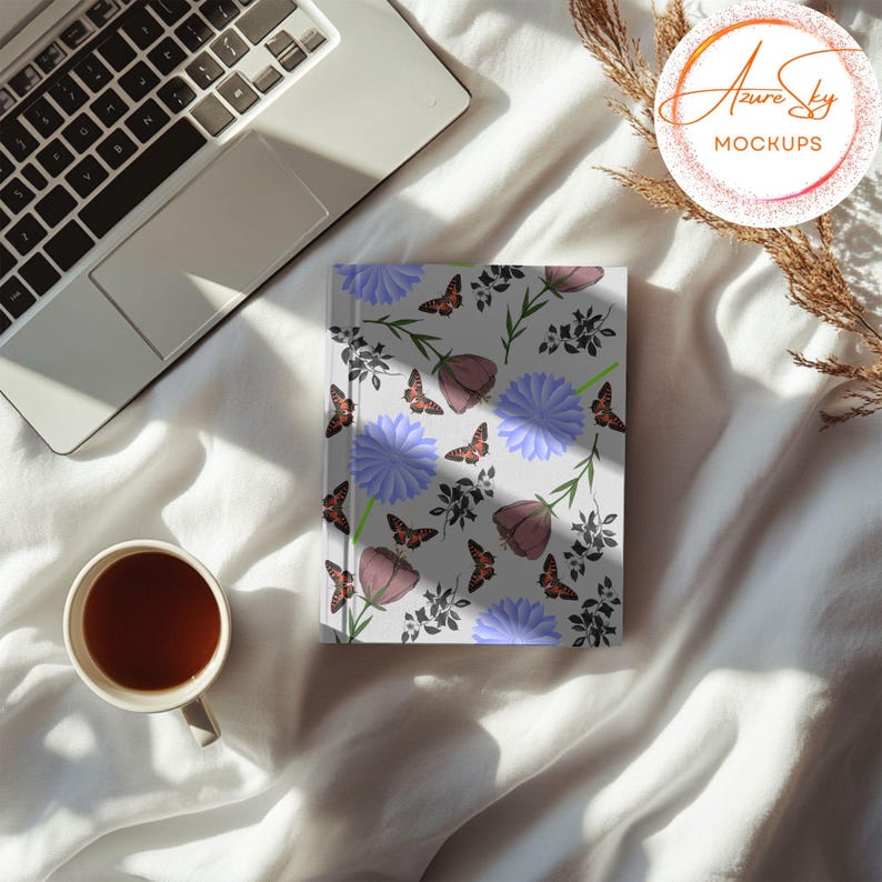 May include: A white notebook with a floral pattern featuring purple flowers, blue flowers, and brown butterflies. The notebook is on a white bedspread with a laptop and a cup of tea.