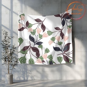 Op de afbeelding: Een witte wandkleed met een bloemenpatroon met groene en bruine bladeren en roze bloemen. De wandkleed hangt aan een grijze betonnen muur.