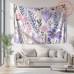 Op de afbeelding: Een witte wandkleed met een bloemenpatroon met roze, paarse en blauwe bloemen. De wandkleed hangt aan een witte muur boven een witte bank.