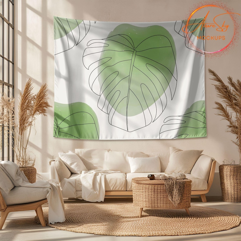 Op de afbeelding: Een witte wandkleed met een groen en wit bladmotief. De wandkleed hangt aan een muur in een woonkamer met een witte bank, een rieten salontafel en een beige kleed.