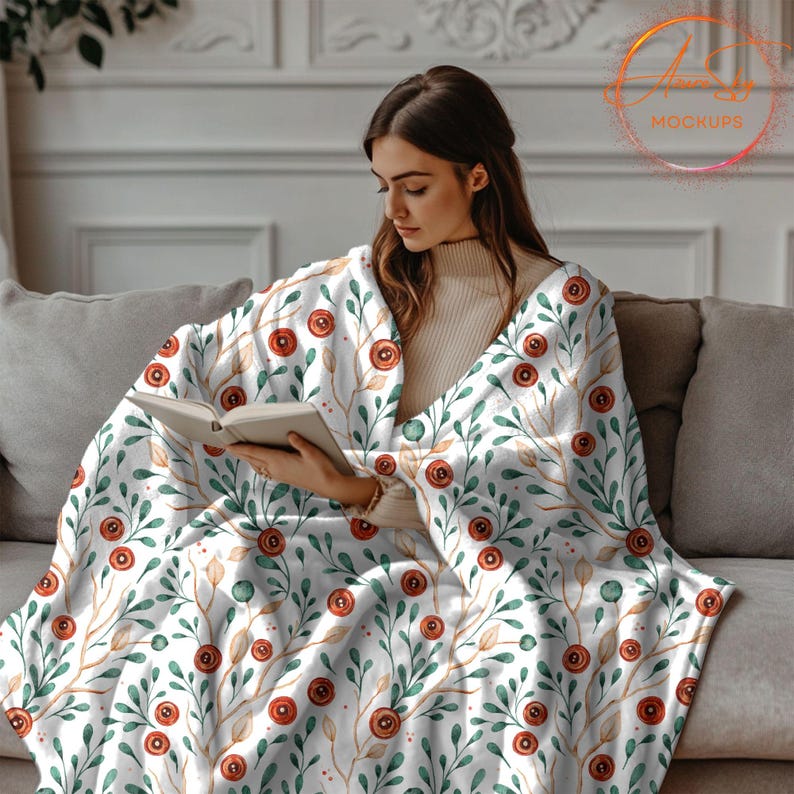 May include: A white throw blanket with a floral pattern featuring red and green watercolor flowers and leaves. The blanket is draped over a couch and a person is sitting on the couch reading a book.