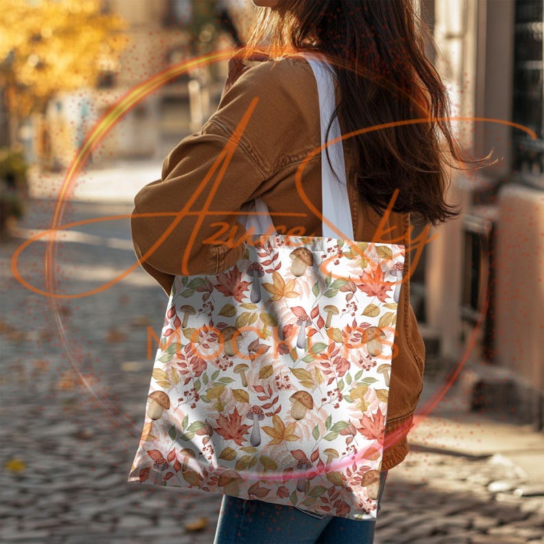 May include: A white tote bag with a autumn-themed design featuring orange, brown, and red leaves, mushrooms, and berries. The bag has a brown leather strap.