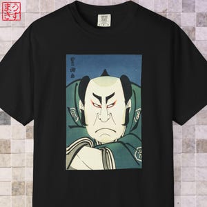 Puede incluir: Camiseta negra con una impresión japonesa Ukiyo-e de un actor de Kabuki. La obra de arte representa a un hombre de rostro severo con maquillaje y vestuario tradicional, con texto japonés encima. La camiseta tiene una etiqueta Comfort Colors.
