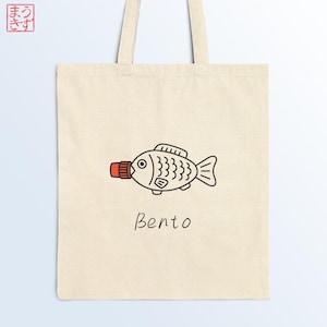 Peut inclure: Un sac fourre-tout beige avec une illustration de poisson noir et rouge, ressemblant à un flacon de sauce soja, et le mot "Bento" en script noir. Le sac a deux anses beiges.