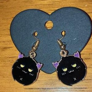 Può includere: Un paio di orecchini pendenti a forma di gatto nero e viola con ganci dorati. Il gatto ha un'espressione scontrosa con occhi gialli e un piccolo corno di diavolo viola su ogni lato della testa.