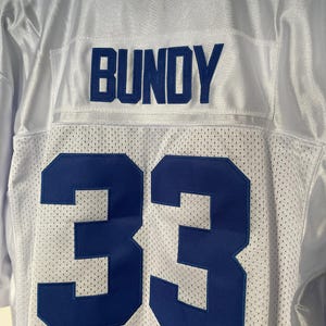 May include: White football jersey with blue lettering. The name "BUNDY" is on the back of the jersey above the number "33".