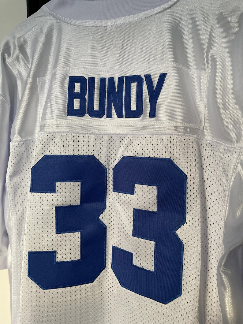 May include: White football jersey with blue lettering. The name "BUNDY" is on the back of the jersey above the number "33".
