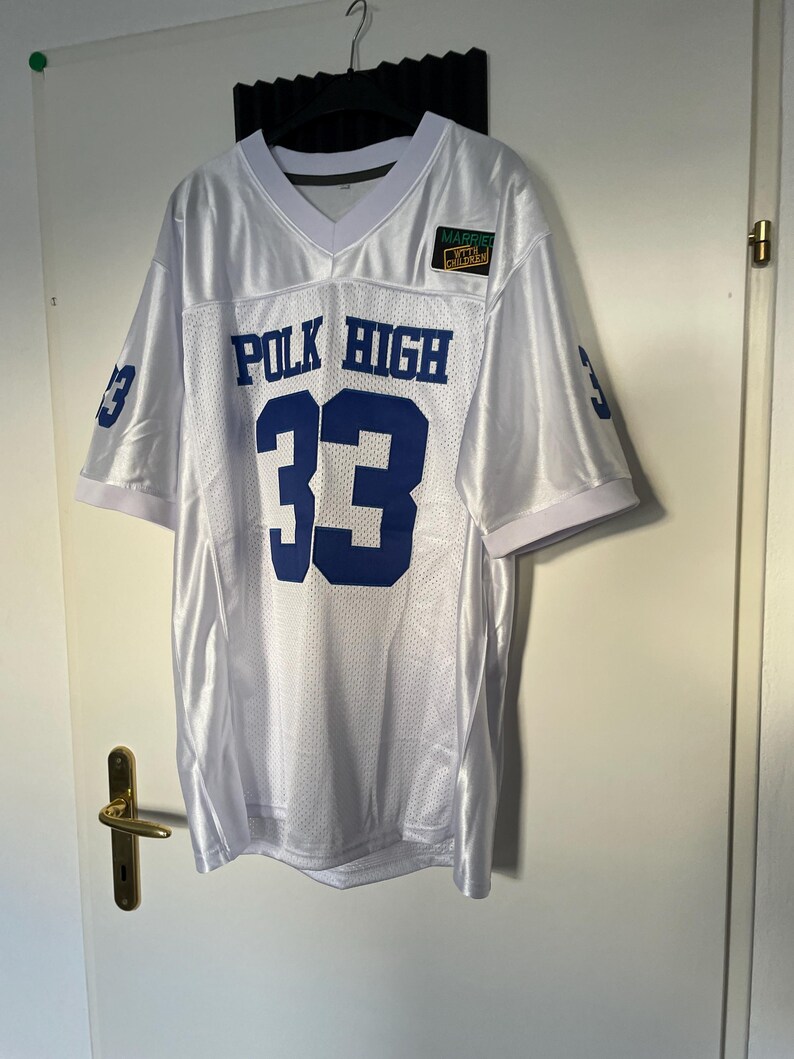 May include: White football jersey with blue lettering that reads "POLK HIGH" and the number "33". A small green patch with the text "MARRIED WITH CHILDREN" is on the right side of the jersey.