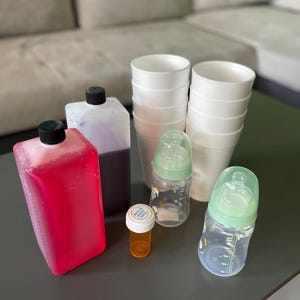 Puede incluir: Un surtido de artículos que incluyen dos biberones con tetinas verdes, un pequeño frasco de pastillas naranja, una pila de vasos de papel blancos y dos botellas rectangulares llenas de líquidos rojos y morados.