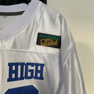 May include: White jersey with blue lettering that says "HIGH" and a green and yellow patch that says "MARRIED W/ CHILDREN".