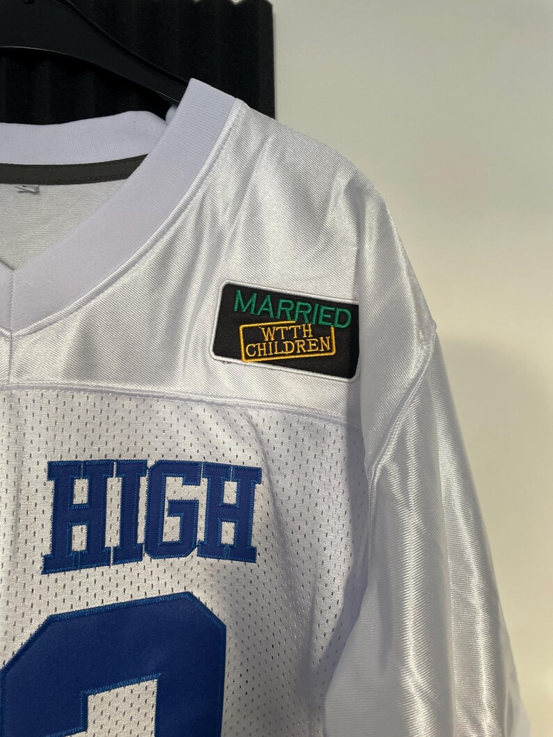 May include: White jersey with blue lettering that says "HIGH" and a green and yellow patch that says "MARRIED W/ CHILDREN".