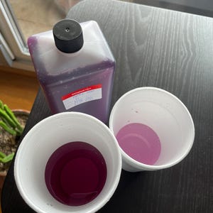 Puede incluir: Una botella de plástico transparente con tapa negra y etiqueta roja, llena de líquido morado, está junto a dos vasos de espuma blancos, cada uno con una porción del mismo líquido. Los vasos y la botella están sobre una superficie oscura.