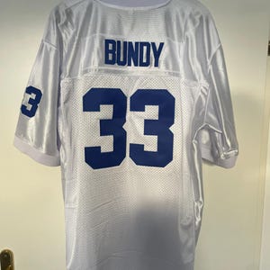 May include: White football jersey with blue lettering on the back. The name "BUNDY" and the number "33" are printed on the back of the jersey.